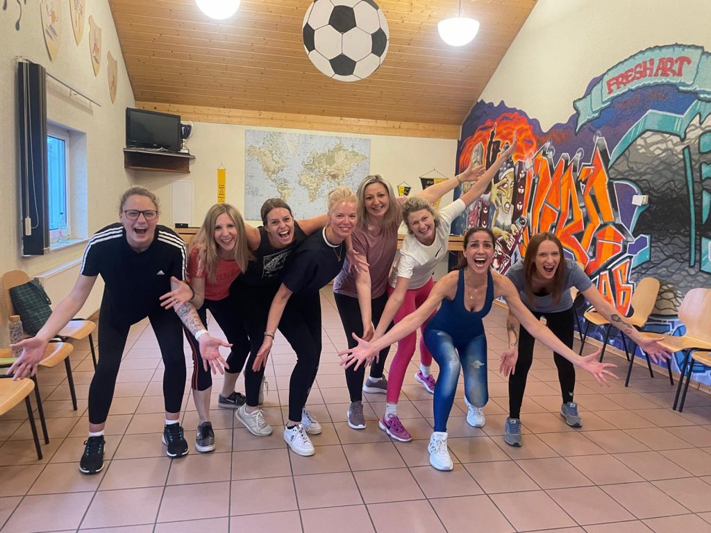 Gruppe von Frauen in einem hellen Raum, die lachend und mit ausgestreckten Armen nach vorne treten – energievolle Szene aus einem Gruppencoaching mit Bewegung und Tanz.
