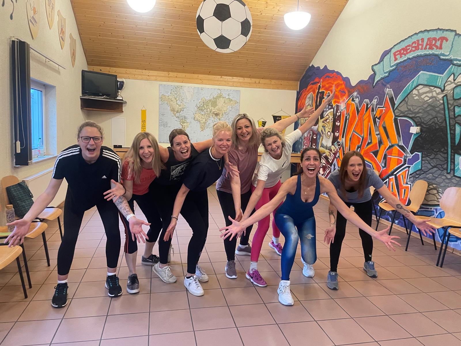 Gruppe von Frauen in einem hellen Raum, die lachend und mit ausgestreckten Armen nach vorne treten – energievolle Szene aus einem Gruppencoaching mit Bewegung und Tanz.