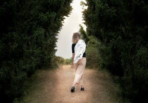 Frau geht allein auf einem schmalen Naturweg zwischen hohen grünen Bäumen und blickt nach vorne in die Ferne