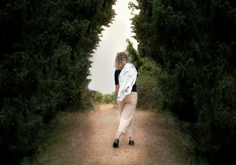 Frau geht allein auf einem schmalen Naturweg zwischen hohen grünen Bäumen und blickt nach vorne in die Ferne