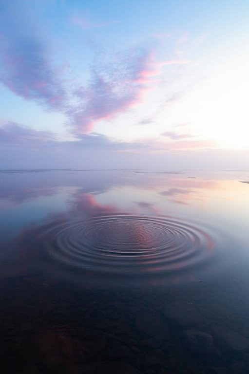 Sanfte Wasseroberfläche mit kreisförmigen Wellen im Vordergrund unter einem pastellfarbenen Himmel in Rosa- und Blautönen.