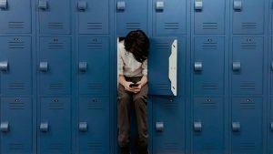 Eine Person sitzt zusammengesunken in einem geöffneten blauen Spind und blickt auf ein Smartphone – umgeben von einer Wand aus Schließfächern.