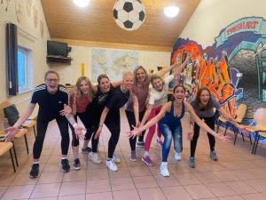 Acht fröhliche Frauen in Sportkleidung stehen lachend und mit ausgebreiteten Armen in einem hellen Raum mit Holzdecke und bunter Graffiti-Wand. Die Gruppe vermittelt Lebensfreude, Gemeinschaft und Spaß an Bewegung.