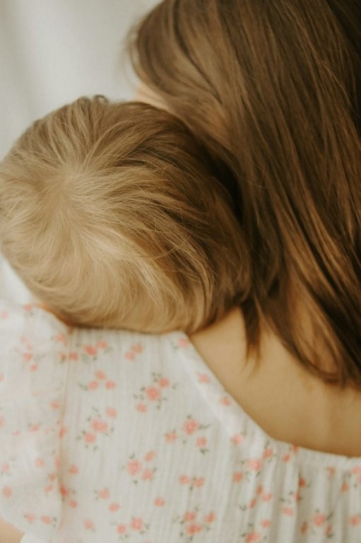 Eine Frau hält ein blondes Kind liebevoll im Arm. Beide lehnen ihre Köpfe aneinander und tragen helle Kleidung mit zartem Blumenmuster. Die Aufnahme vermittelt Geborgenheit, Nähe und Verbundenheit.