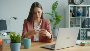 Junge Frau sitzt an einem Schreibtisch im Büro, blickt konzentriert auf ihr Smartphone. Vor ihr stehen ein Laptop, eine Tasse und eine Zimmerpflanze in einer hellen, modernen Arbeitsumgebung.