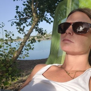 Eine Frau liegt entspannt auf einer grünen Liege am Ufer eines ruhigen Sees. Sie trägt eine Sonnenbrille und genießt die warmen Sonnenstrahlen, während Bäume im Vordergrund und das stille Wasser im Hintergrund eine friedliche Atmosphäre schaffen.